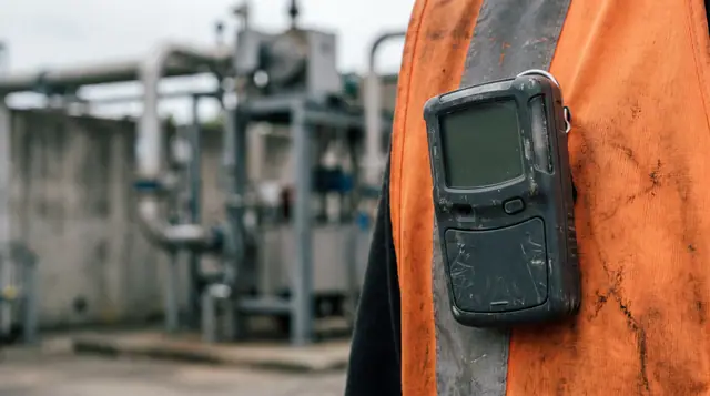 Personal gas detector clipped to a worker's safety vest, blank display visible, with a blurred